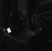 A black and white low-light photograph showing two individuals in tactical gear operating in a dark environment. One person is holding what appears to be a firearm with an attached illuminated device, possibly a weapon-mounted light or targeting system. Partial text visible on one individual's vest suggests law enforcement or tactical unit affiliation.