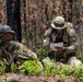 Two military personnel in multicam camouflage uniforms are seated on the ground in a forested area, appearing to review a document. The soldier on the right has a visible 'EOD' (Explosive Ordnance Disposal) patch and is holding a radio while reading a white paper. The surrounding environment shows signs of a previous fire, with charred tree trunks and dead foliage.