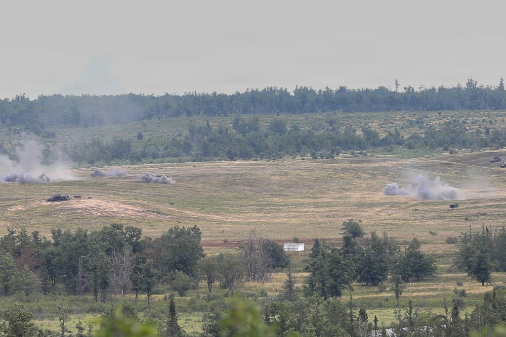 A wide-angle view of an open military training range or exercise area showing multiple smoke plumes rising from what appear to be detonations or firing impacts across a grassy field. Several armored vehicles are faintly visible at distance amid the smoke. The scene is observed from an elevated vantage point across a treeline foreground.