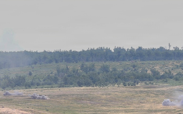 A wide-angle view of an open military training range or exercise area showing multiple smoke plumes rising from what appear to be detonations or firing impacts across a grassy field. Several armored vehicles are faintly visible at distance amid the smoke. The scene is observed from an elevated vantage point across a treeline foreground.