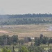 A wide-angle view of an open military training range or exercise area showing multiple smoke plumes rising from what appear to be detonations or firing impacts across a grassy field. Several armored vehicles are faintly visible at distance amid the smoke. The scene is observed from an elevated vantage point across a treeline foreground.