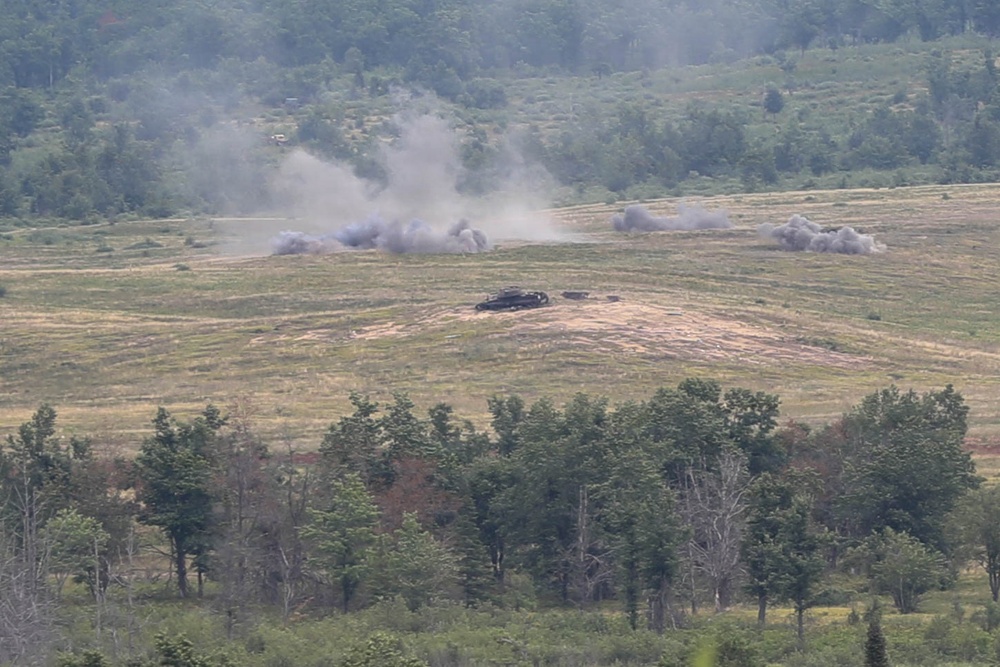 The image shows a military training exercise or combat scenario taking place on an open field surrounded by forested hills. Multiple explosion or smoke plumes are visible across the field, with at least one armored vehicle visible in the center of the impact zone. The scene appears to be captured from an elevated observation point at a considerable distance.