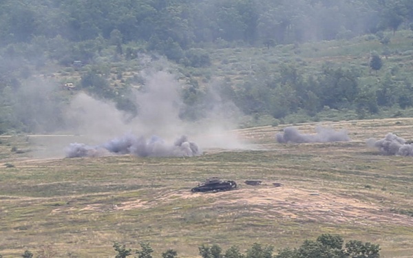 The image shows a military training exercise or combat scenario taking place on an open field surrounded by forested hills. Multiple explosion or smoke plumes are visible across the field, with at least one armored vehicle visible in the center of the impact zone. The scene appears to be captured from an elevated observation point at a considerable distance.