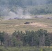 The image shows a military training exercise or combat scenario taking place on an open field surrounded by forested hills. Multiple explosion or smoke plumes are visible across the field, with at least one armored vehicle visible in the center of the impact zone. The scene appears to be captured from an elevated observation point at a considerable distance.
