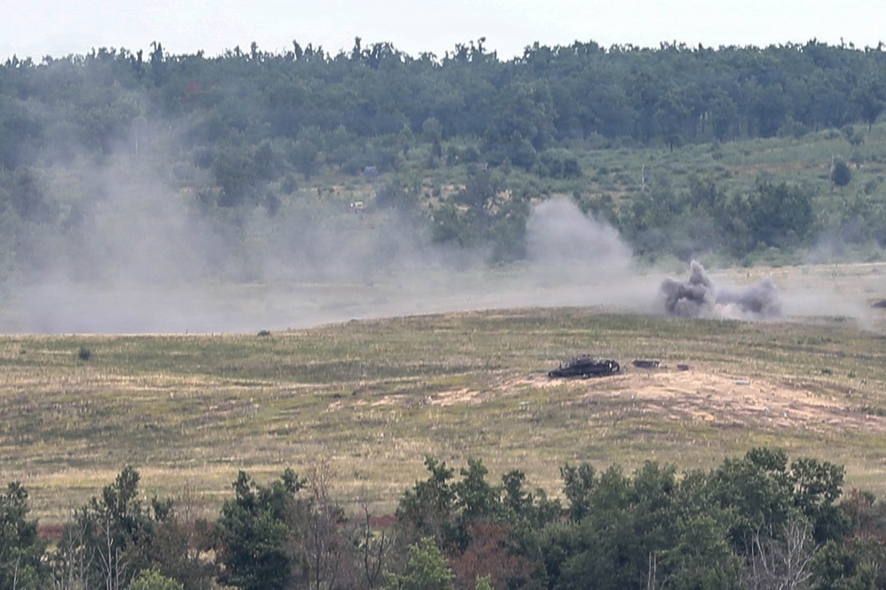 The image shows an open field or training range with what appears to be an explosion or detonation occurring in the mid-ground. A dark, destroyed or burning vehicle is visible on the ground, with large plumes of gray smoke rising from the blast area. The scene is consistent with a military live-fire exercise or weapons testing on a range.