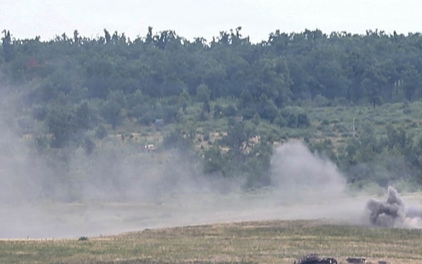 The image shows an open field or training range with what appears to be an explosion or detonation occurring in the mid-ground. A dark, destroyed or burning vehicle is visible on the ground, with large plumes of gray smoke rising from the blast area. The scene is consistent with a military live-fire exercise or weapons testing on a range.