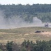 The image shows an open field or training range with what appears to be an explosion or detonation occurring in the mid-ground. A dark, destroyed or burning vehicle is visible on the ground, with large plumes of gray smoke rising from the blast area. The scene is consistent with a military live-fire exercise or weapons testing on a range.