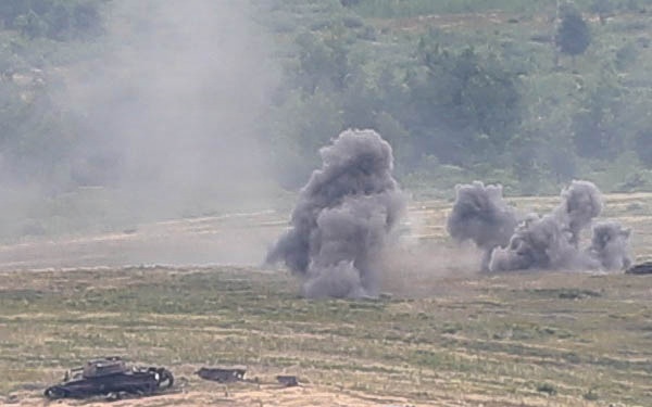 The image shows an open field or training range with multiple explosion or detonation plumes rising from the ground. At least one armored vehicle or military equipment is visible in the foreground, appearing damaged or stationary. The scene is consistent with a military exercise, live-fire training, or combat engagement.