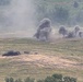 The image shows an open field or training range with multiple explosion or detonation plumes rising from the ground. At least one armored vehicle or military equipment is visible in the foreground, appearing damaged or stationary. The scene is consistent with a military exercise, live-fire training, or combat engagement.