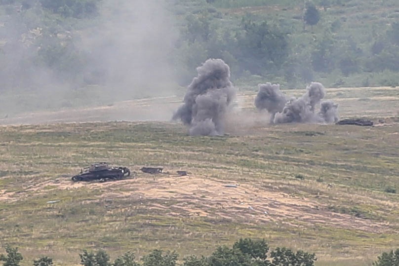 The image shows an open field or training range with multiple explosion or detonation plumes rising from the ground. At least one armored vehicle or military equipment is visible in the foreground, appearing damaged or stationary. The scene is consistent with a military exercise, live-fire training, or combat engagement.