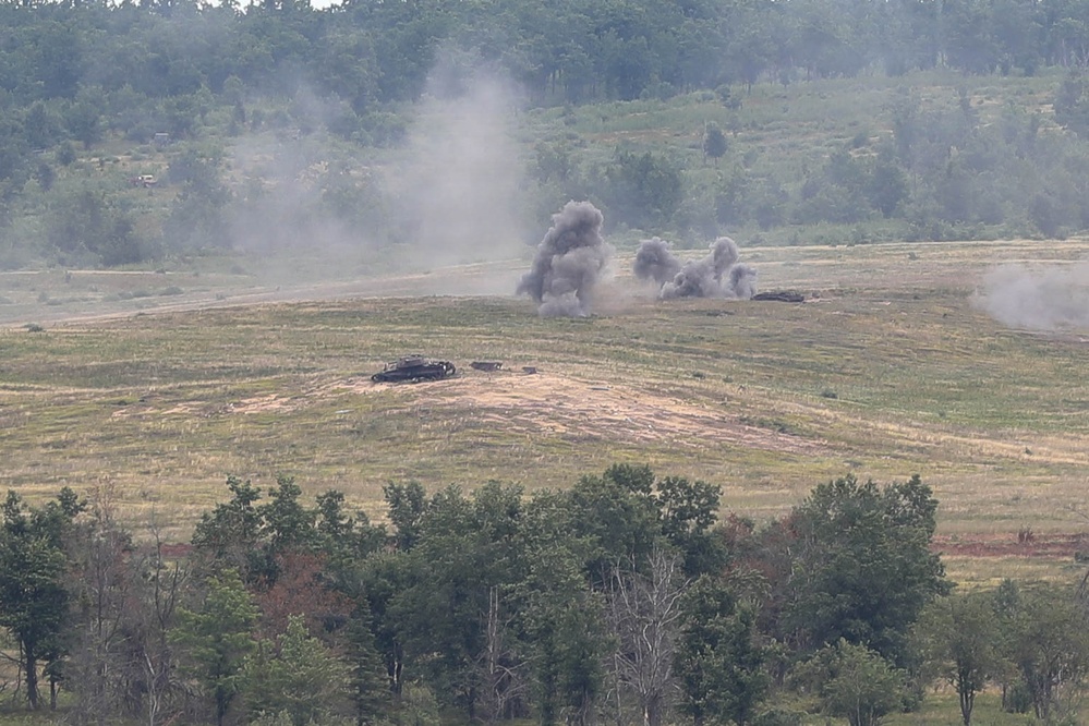 The image shows a military training or live-fire exercise on an open range with explosions and smoke visible in the mid-ground. At least one armored vehicle or target is visible on the terrain, with multiple detonations producing dark smoke columns. The scene is set in a hilly, partially wooded landscape consistent with a military training installation.