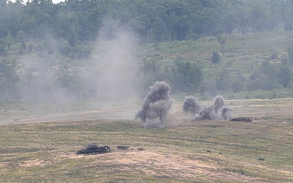 The image shows a military training or live-fire exercise on an open range with explosions and smoke visible in the mid-ground. At least one armored vehicle or target is visible on the terrain, with multiple detonations producing dark smoke columns. The scene is set in a hilly, partially wooded landscape consistent with a military training installation.