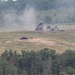 The image shows a military training or live-fire exercise on an open range with explosions and smoke visible in the mid-ground. At least one armored vehicle or target is visible on the terrain, with multiple detonations producing dark smoke columns. The scene is set in a hilly, partially wooded landscape consistent with a military training installation.