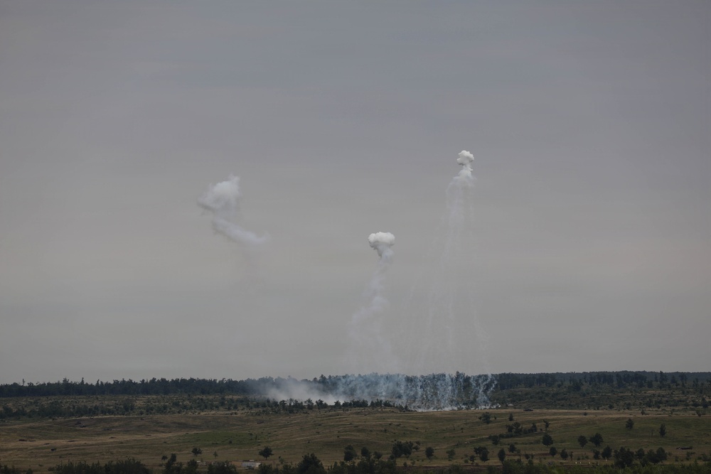 The image shows a distant landscape with multiple white smoke plumes rising from a treeline into an overcast gray sky. Three distinct smoke columns are visible at varying heights, with a broader smoke dispersion pattern along the ground near the treeline. The scene is consistent with artillery, mortar, or smoke munition activity on what appears to be open terrain.