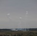 The image shows a distant landscape with multiple white smoke plumes rising from a treeline into an overcast gray sky. Three distinct smoke columns are visible at varying heights, with a broader smoke dispersion pattern along the ground near the treeline. The scene is consistent with artillery, mortar, or smoke munition activity on what appears to be open terrain.