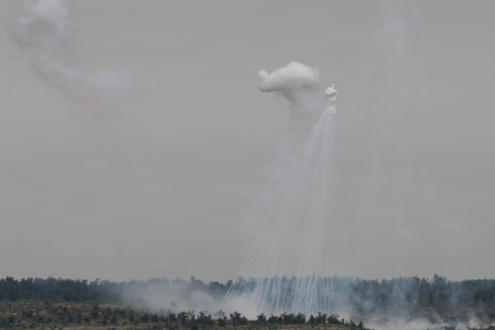 The image shows an aerial burst producing multiple white streaking trails descending from a central explosion point in the sky. A large white cloud of smoke or dispersed material is visible at the apex, with numerous thin white lines radiating downward toward a treeline. Additional smoke is visible at ground level and to the left of the frame.