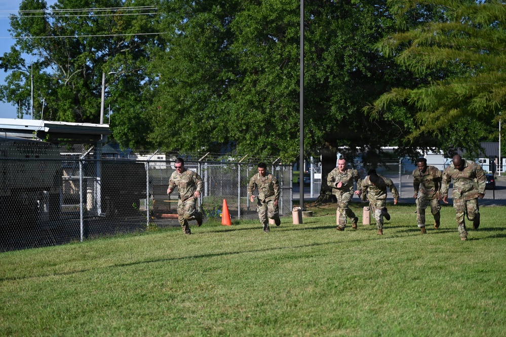 A group of approximately six military personnel in camouflage uniforms are running across a grassy field during what appears to be a physical training exercise. They are moving in a line formation past an orange traffic cone marker, suggesting a timed or competitive drill. The setting appears to be a military installation or base, indicated by chain-link fencing and institutional buildings in the background.