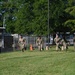 A group of approximately six military personnel in camouflage uniforms are running across a grassy field during what appears to be a physical training exercise. They are moving in a line formation past an orange traffic cone marker, suggesting a timed or competitive drill. The setting appears to be a military installation or base, indicated by chain-link fencing and institutional buildings in the background.