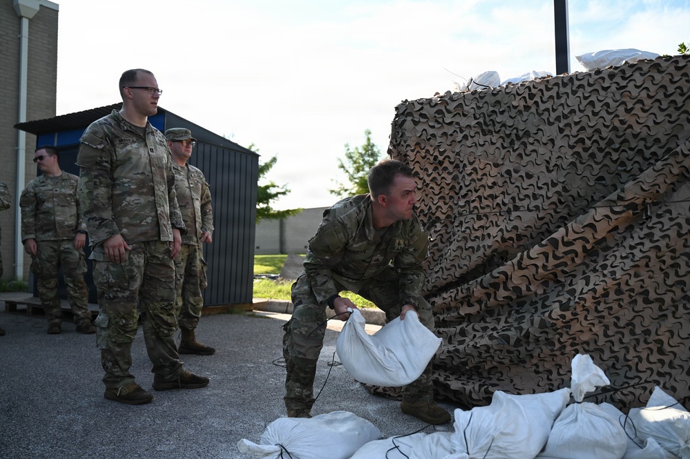 Military personnel in MultiCam/OCP camouflage uniforms are engaged in what appears to be flood preparedness or defensive operations involving sandbags and camouflage netting. One soldier is actively placing or arranging sandbags near a large stack of camouflage netting, while three other soldiers observe. The setting appears to be a military installation or base facility.