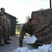 Military personnel in MultiCam/OCP camouflage uniforms are engaged in what appears to be flood preparedness or defensive operations involving sandbags and camouflage netting. One soldier is actively placing or arranging sandbags near a large stack of camouflage netting, while three other soldiers observe. The setting appears to be a military installation or base facility.