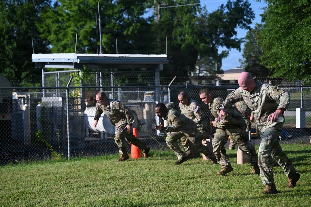 A group of military personnel in camouflage uniforms are performing a physical training exercise on a grass field. They appear to be in a starting sprint position or conducting a running drill in a line formation. The setting appears to be a military installation based on the chain-link fence with barbed wire and surrounding infrastructure.