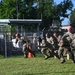 A group of military personnel in camouflage uniforms are performing a physical training exercise on a grass field. They appear to be in a starting sprint position or conducting a running drill in a line formation. The setting appears to be a military installation based on the chain-link fence with barbed wire and surrounding infrastructure.