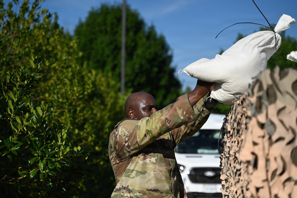 A military service member in OCP (Operational Camouflage Pattern) uniform is lifting a white sandbag overhead, placing it onto a stack covered with camouflage netting. The activity appears to be flood control or defensive barrier construction. The setting is outdoors with green trees, blue sky, and a white vehicle visible in the background.