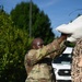 A military service member in OCP (Operational Camouflage Pattern) uniform is lifting a white sandbag overhead, placing it onto a stack covered with camouflage netting. The activity appears to be flood control or defensive barrier construction. The setting is outdoors with green trees, blue sky, and a white vehicle visible in the background.