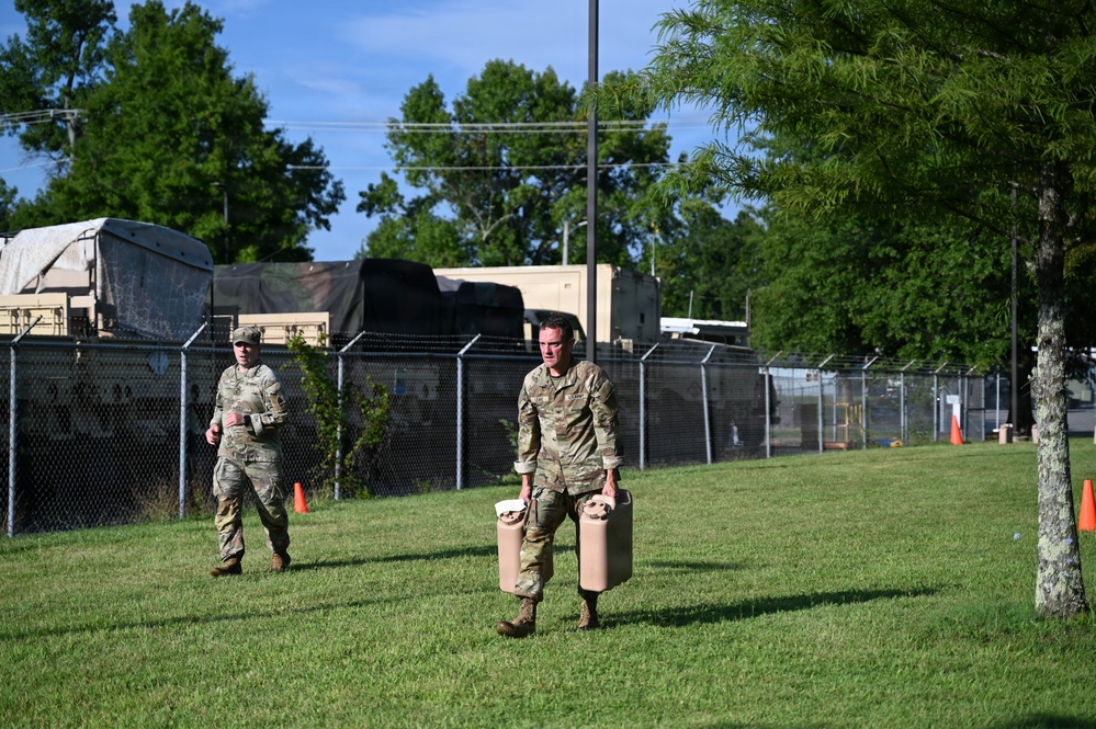 Two military personnel in OCP (Operational Camouflage Pattern) uniforms are walking across a grassy area within what appears to be a military installation or base. The soldier in the foreground is carrying two tan/beige jerry cans or similar containers. Military vehicles and a chain-link fence with barbed wire are visible in the background.