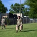 Two military personnel in OCP (Operational Camouflage Pattern) uniforms are walking across a grassy area within what appears to be a military installation or base. The soldier in the foreground is carrying two tan/beige jerry cans or similar containers. Military vehicles and a chain-link fence with barbed wire are visible in the background.