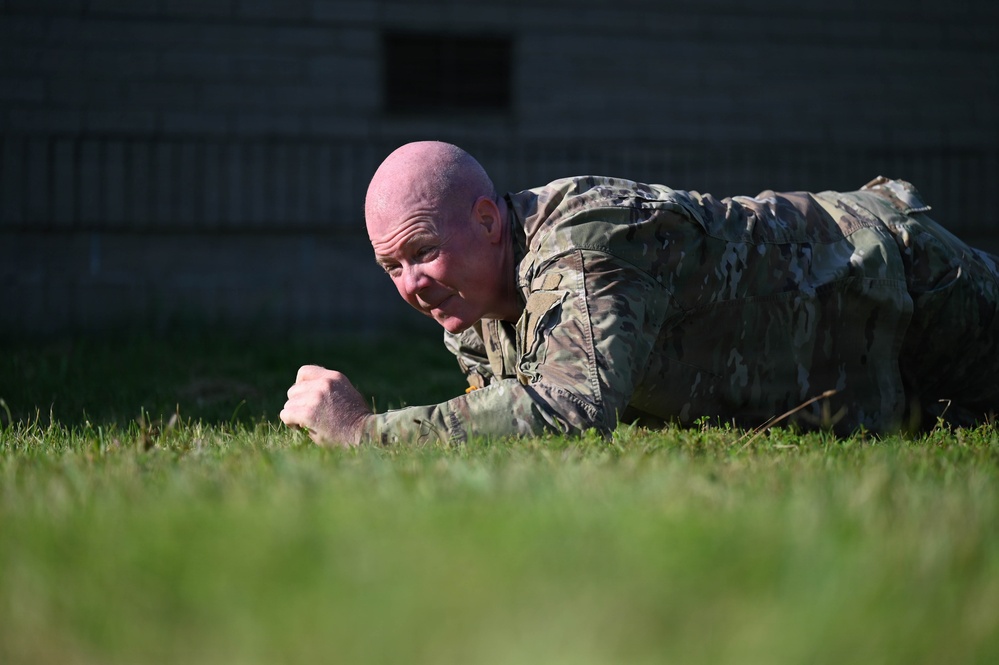 A bald male individual wearing multicam/OCP camouflage military uniform is performing a prone plank exercise on a grass field. He is lying face-down with fists clenched on the ground, supporting his body weight in a low plank position. A building structure is visible in the blurred background.