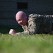 A bald male individual wearing multicam/OCP camouflage military uniform is performing a prone plank exercise on a grass field. He is lying face-down with fists clenched on the ground, supporting his body weight in a low plank position. A building structure is visible in the blurred background.