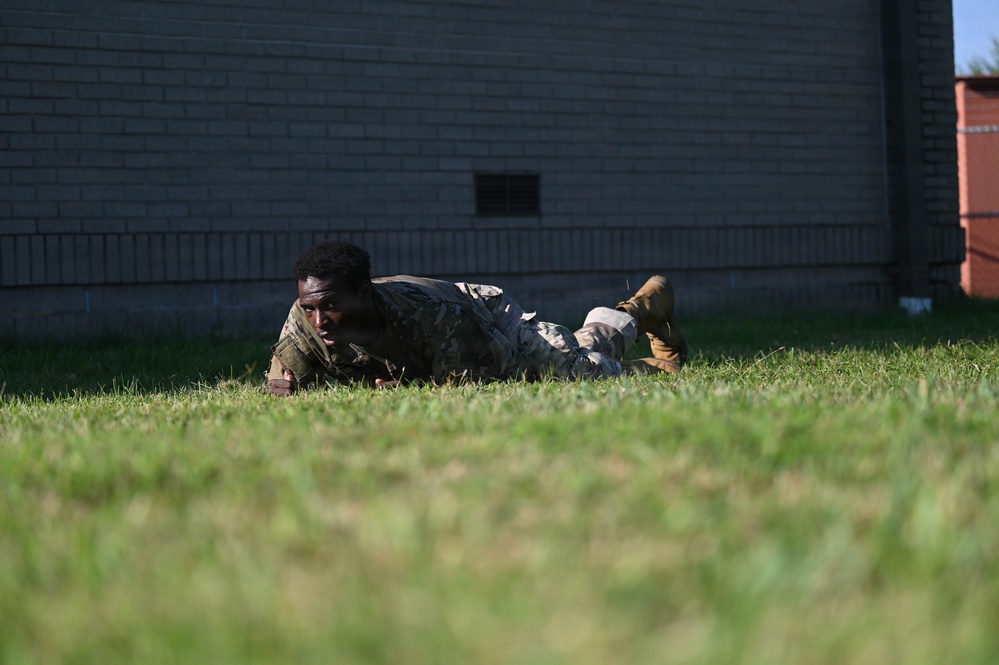 A soldier in camouflage uniform is performing a low crawl maneuver on a grass field near a brick building. The individual appears to be in a training exercise, executing a tactical ground movement technique. The photo is taken from a low angle at ground level, emphasizing the crawling motion.
