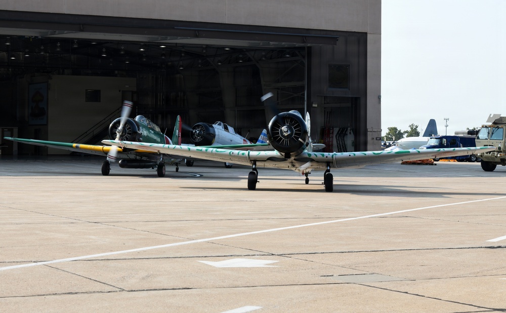The image shows multiple vintage/warbird propeller aircraft parked on a concrete flight line apron in front of a large military-style hangar. The foreground aircraft displays a distinctive green and yellow camouflage paint scheme with a radial engine, while additional similar aircraft are visible near the open hangar doors. Ground support vehicles and equipment are visible to the right side of the frame.