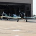 The image shows multiple vintage/warbird propeller aircraft parked on a concrete flight line apron in front of a large military-style hangar. The foreground aircraft displays a distinctive green and yellow camouflage paint scheme with a radial engine, while additional similar aircraft are visible near the open hangar doors. Ground support vehicles and equipment are visible to the right side of the frame.