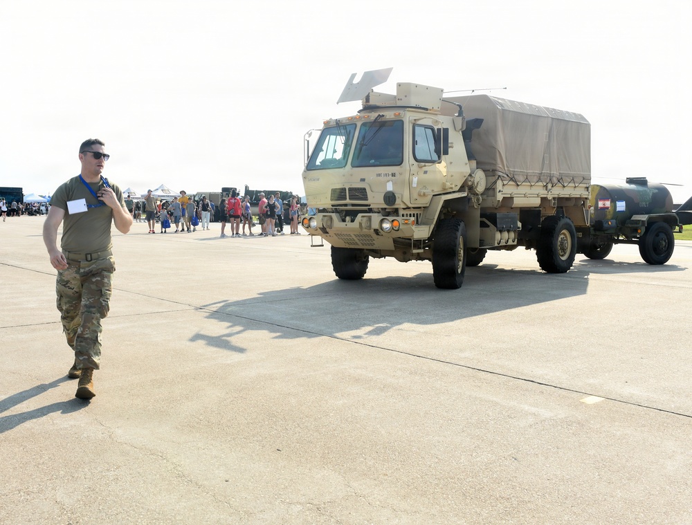 A military service member in camouflage uniform walks across a tarmac or airfield apron during what appears to be a public air show or military open house event. A tan/desert-colored military tactical truck (LMTV or similar Medium Tactical Vehicle) towing a trailer is parked nearby. Civilians and families can be seen gathered in the background, consistent with a public display event.