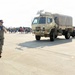 A military service member in camouflage uniform walks across a tarmac or airfield apron during what appears to be a public air show or military open house event. A tan/desert-colored military tactical truck (LMTV or similar Medium Tactical Vehicle) towing a trailer is parked nearby. Civilians and families can be seen gathered in the background, consistent with a public display event.