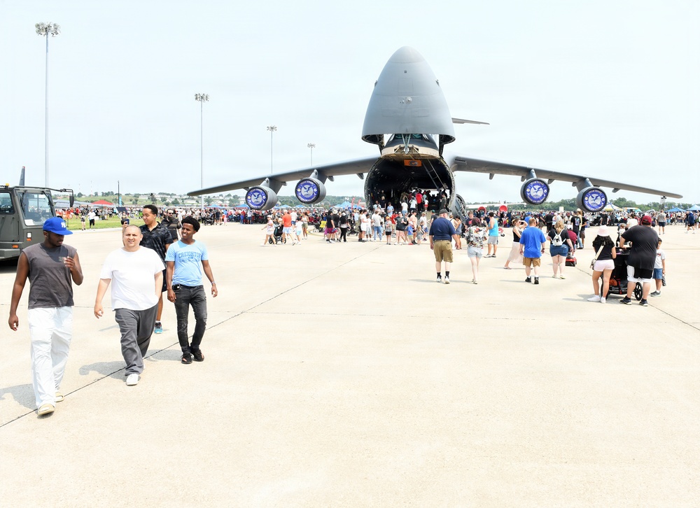 A large military air show event on an airfield tarmac featuring a massive C-5 Galaxy military transport aircraft with its nose cargo door open. Hundreds of civilians are gathered around and walking through the aircraft as part of a public static display. The event appears to be a well-attended open house or air show at a U.S. Air Force base.