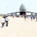 A large military air show event on an airfield tarmac featuring a massive C-5 Galaxy military transport aircraft with its nose cargo door open. Hundreds of civilians are gathered around and walking through the aircraft as part of a public static display. The event appears to be a well-attended open house or air show at a U.S. Air Force base.