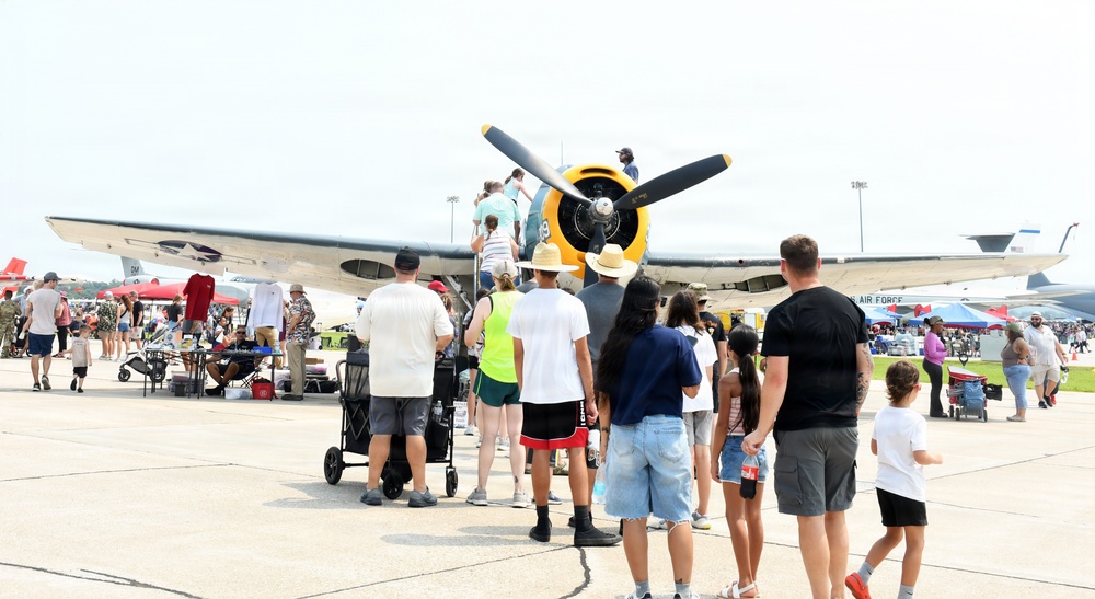 A public airshow event at a military or civilian airfield where crowds of people are gathered around a vintage propeller aircraft with a distinctive yellow engine cowling. Visitors are closely inspecting the aircraft, with some individuals climbing onto or near the plane for a closer look. Multiple aircraft and vendor tents are visible in the background, indicating a large-scale aviation exhibition.