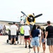 A public airshow event at a military or civilian airfield where crowds of people are gathered around a vintage propeller aircraft with a distinctive yellow engine cowling. Visitors are closely inspecting the aircraft, with some individuals climbing onto or near the plane for a closer look. Multiple aircraft and vendor tents are visible in the background, indicating a large-scale aviation exhibition.