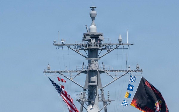 A U.S. Navy Arleigh Burke-class guided-missile destroyer is shown in a ceremonial homecoming or commissioning event, viewed from a bow-on perspective. Sailors in dress white uniforms are manning the rails along the bow, and multiple flags including the American flag and a black ship's commissioning/battle flag are flying from the mast. A harbor tugboat in orange and yellow is visible alongside the starboard bow.