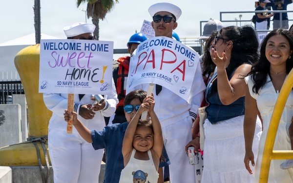 A military homecoming event at what appears to be a naval pier, likely in San Diego based on the 'SD Navy League' branding on the signs. Families and children are gathered to welcome returning Navy personnel, holding signs reading 'Welcome Home Sweet Home' and 'Welcome Home Papa'. The scene captures an emotional and joyful reunion atmosphere with Navy sailors in white dress uniforms present.
