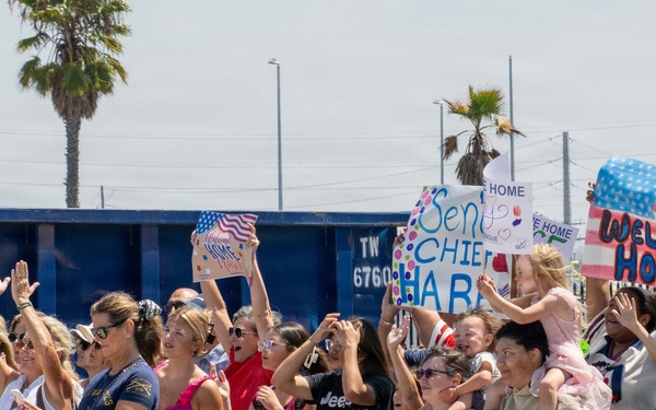 A crowd of people, predominantly women and children, are gathered outdoors for a military homecoming event. They are waving, cheering, and holding handmade signs with messages such as 'Welcome Home,' 'Hero's Welcome Home,' and 'Senior Chief Hab[er].' The setting appears to be a naval or military base, likely in Southern California based on the palm trees and coastal environment.