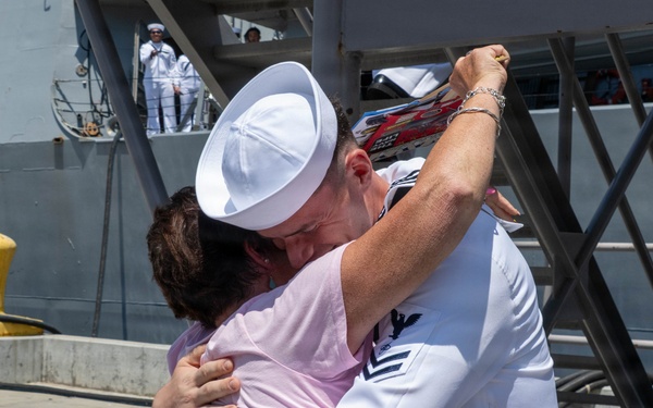 A U.S. Navy sailor in dress white uniform embraces a civilian woman on a pier next to a naval vessel. The emotional reunion appears to be a homecoming event, with additional sailors visible on the ship's deck in the background. The scene takes place at a naval port or dock facility.