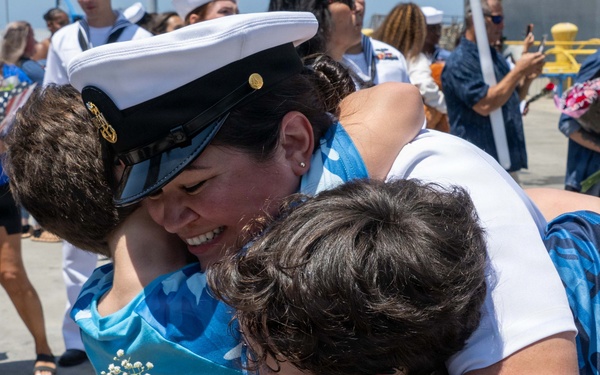 A U.S. Navy sailor in dress whites is reuniting with family members — appearing to embrace at least two people simultaneously, including a child — at what appears to be a homecoming event on a pier or dock. The sailor is smiling broadly and holding red roses with baby's breath. The background shows other sailors and civilians gathered in a similar celebratory context.