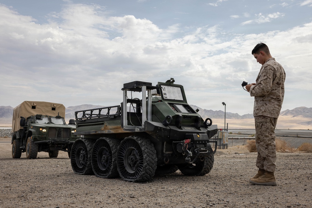 A U.S. Marine in desert camouflage (MARPAT) uniform operates what appears to be an unmanned ground vehicle (UGV) using a handheld controller in an arid desert environment. The robotic vehicle is a large 8-wheeled autonomous ground platform with airless/non-pneumatic tires, likely used for logistics or resupply missions. Additional military vehicles are visible in the background, suggesting a field exercise or technology demonstration.