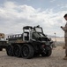 A U.S. Marine in desert camouflage (MARPAT) uniform operates what appears to be an unmanned ground vehicle (UGV) using a handheld controller in an arid desert environment. The robotic vehicle is a large 8-wheeled autonomous ground platform with airless/non-pneumatic tires, likely used for logistics or resupply missions. Additional military vehicles are visible in the background, suggesting a field exercise or technology demonstration.