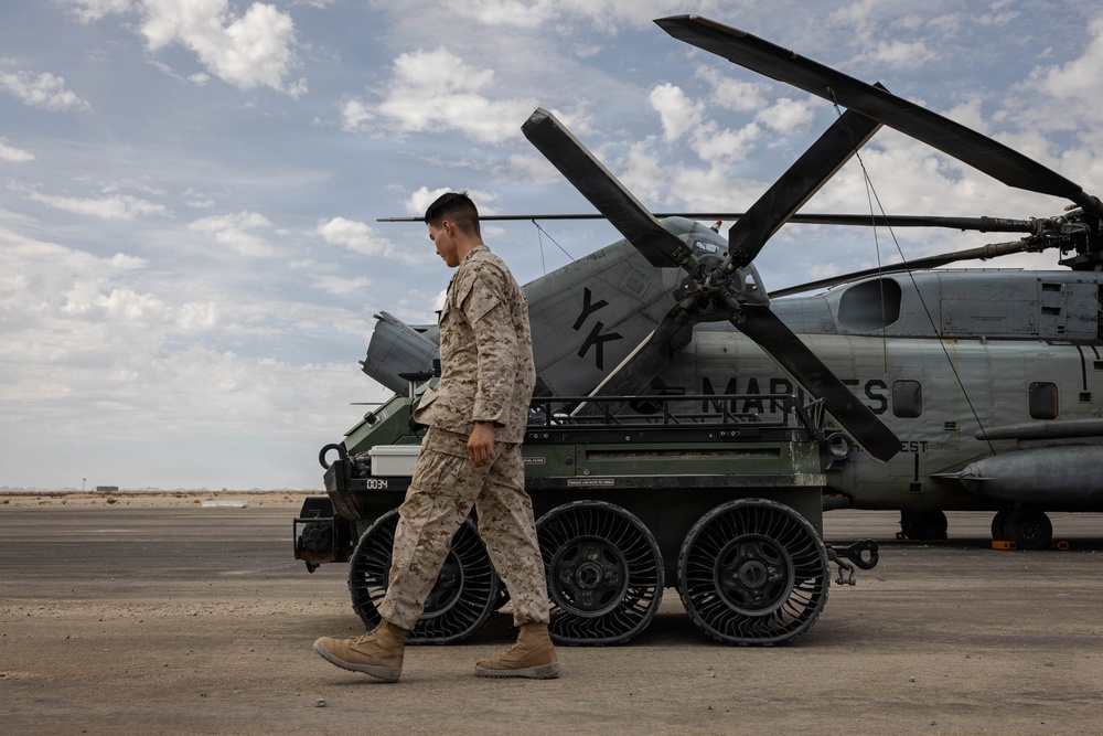 A U.S. Marine in desert camouflage (MARPAT) uniform walks across a tarmac or airfield in what appears to be an arid environment. Behind him is a ground vehicle with distinctive airless/non-pneumatic tires and a large military helicopter marked 'MARINES' with folded rotor blades. The setting suggests a forward operating base or military airfield in a desert region.