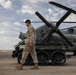 A U.S. Marine in desert camouflage (MARPAT) uniform walks across a tarmac or airfield in what appears to be an arid environment. Behind him is a ground vehicle with distinctive airless/non-pneumatic tires and a large military helicopter marked 'MARINES' with folded rotor blades. The setting suggests a forward operating base or military airfield in a desert region.